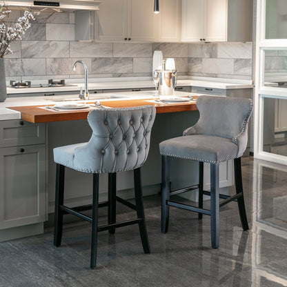 Two Contemporary Velvet Upholstered Wing-Back Barstools with button tufting and chrome nailhead trim sit at a modern kitchen island with a wooden countertop, surrounded by white cabinetry and a gray marble tile backsplash.