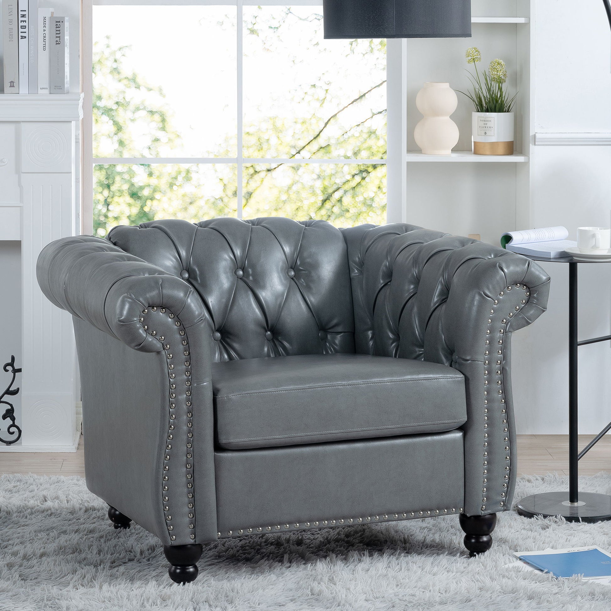 The 1 Seater Sofa For Living Room features gray tufted leather, rolled arms, and nailhead trim, set in a bright space with white shelves, decor accents, and a large window revealing greenery outside.