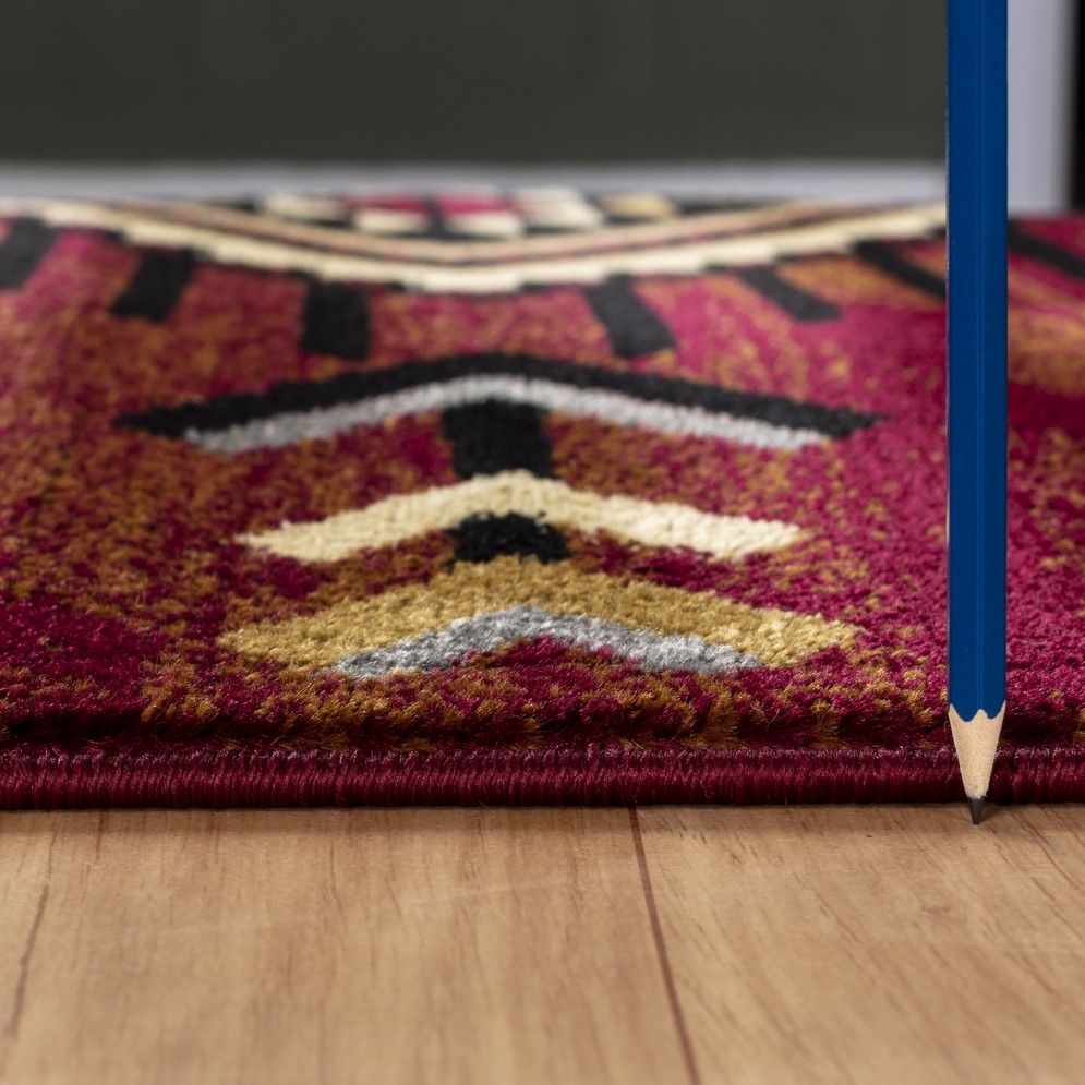 A close-up side view of the Tribes - GC_YLS4002 Southwest Area Rug, thin and patterned, on a wooden floor with a blue pencil standing vertically beside its edge.