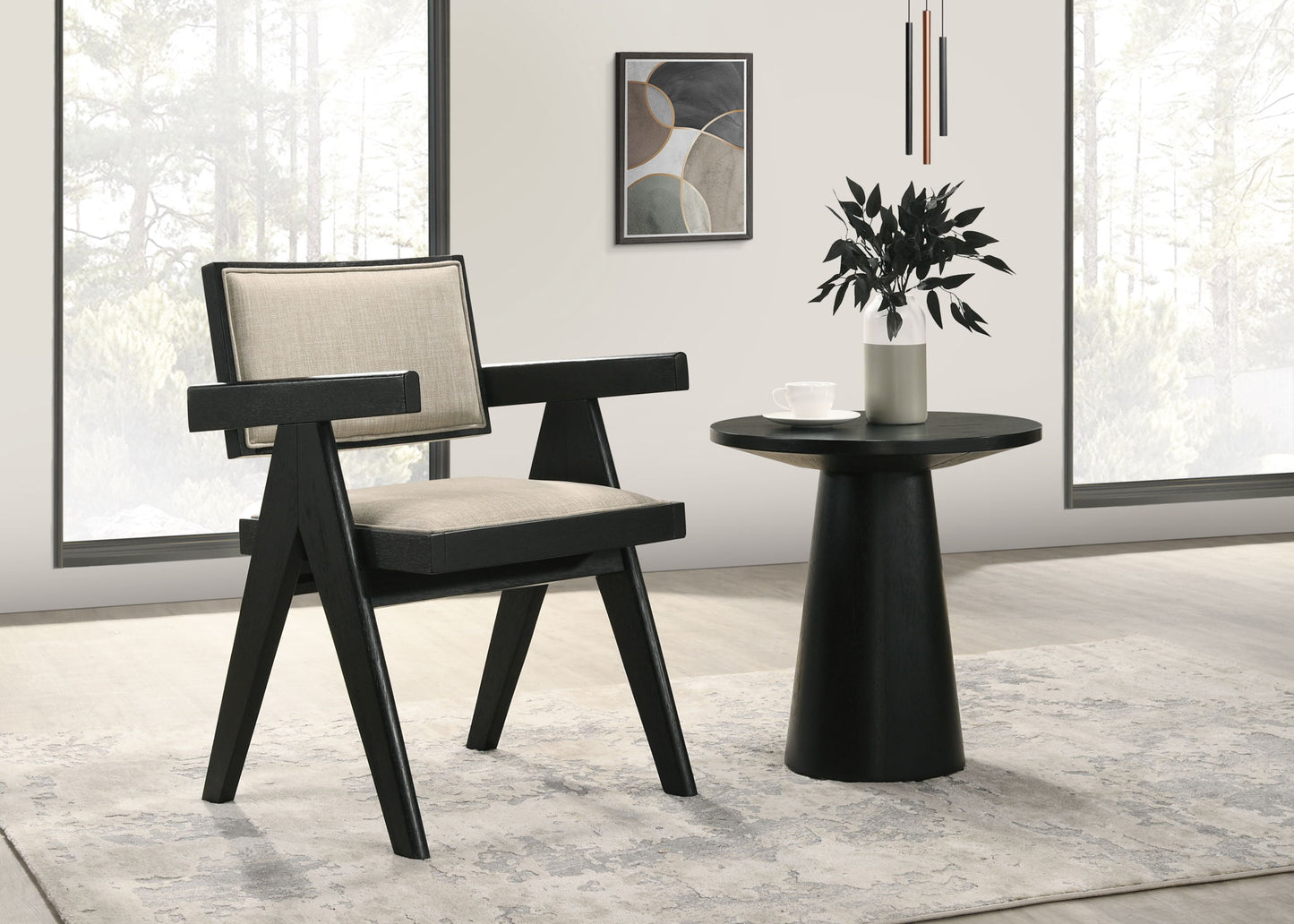 A modern living room featuring the Jasper 3 Piece Arm Chairs and Console Table set in Ebony Black, with beige cushions, accented by a plant and cup on the table, abstract art on the wall, and large windows in the background.