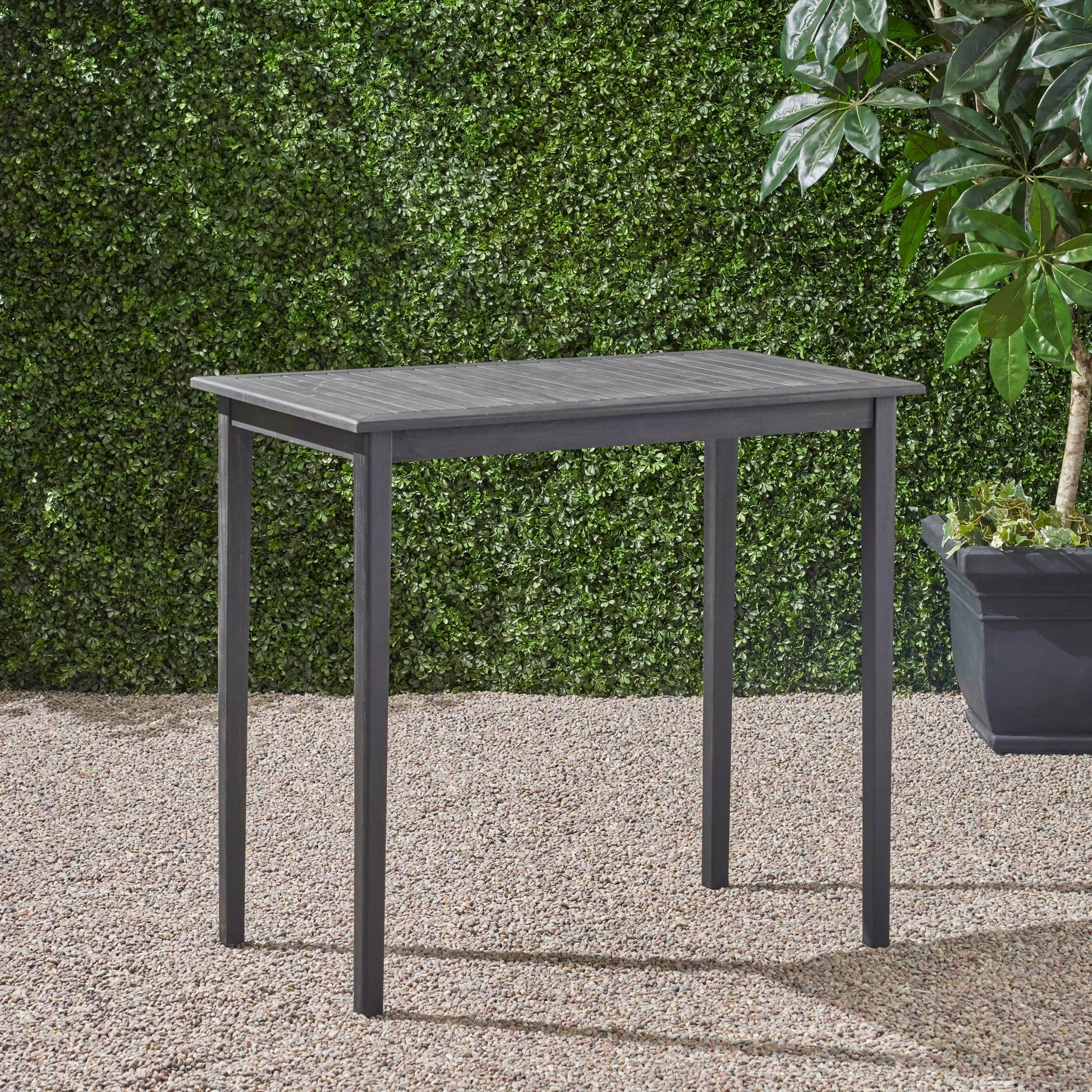 The Outdoor Minimalist Acacia Wood Rectangle Bar Table, featuring a simple rectangular design and straight legs in black, stands on a gravel surface outdoors beside a leafy potted plant and a green hedge wall.