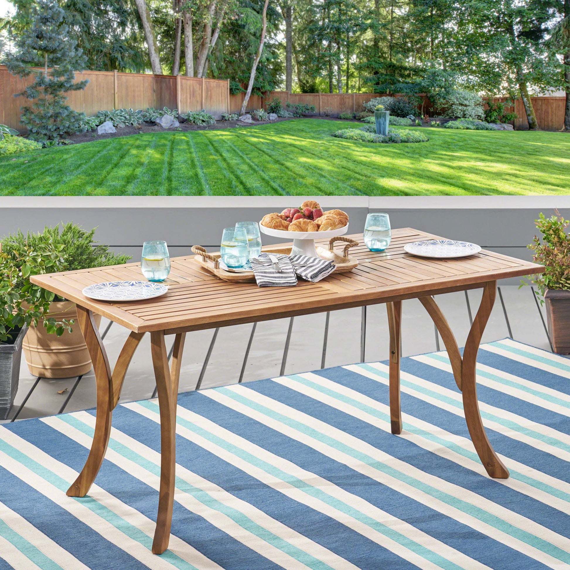 An Outdoor Rectangular Table With Curved Legs And Perpendicular Wooden Slats Table Top sits on a striped blue and white rug, set with plates, water glasses, napkins, and a tray of pastries and fruit on a patio overlooking a landscaped lawn.