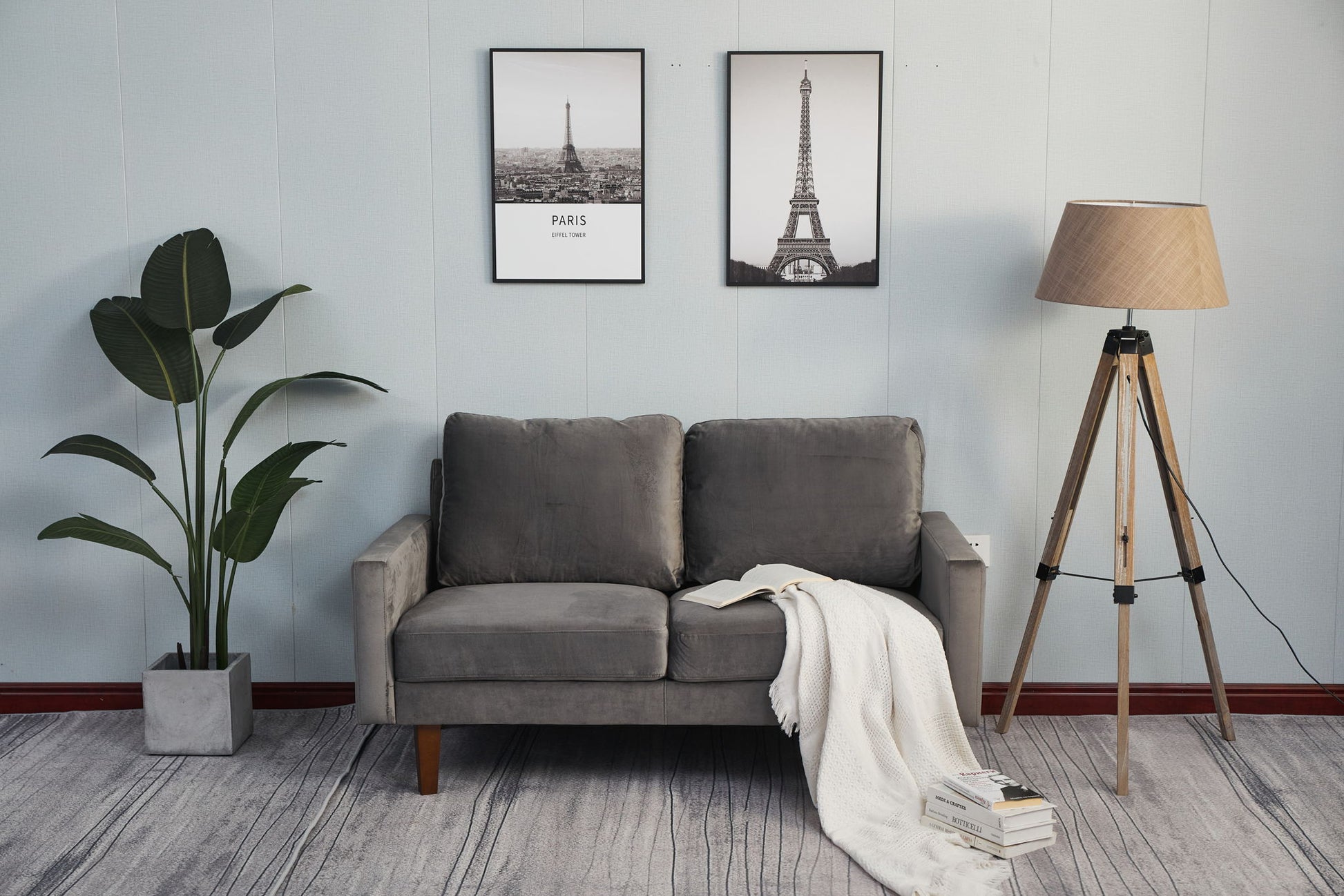 A modern living room features the Comfortable Loveseat With Durable Construction, a white blanket, open book, two Eiffel Tower prints, tall floor lamp, potted plant, and stacked books on a light-striped rug.