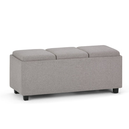 The Avalon Upholstered Storage Ottoman features a rectangular gray design, three cushioned sections, and short black legs, shown against a white background.