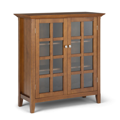 The Acadian - Medium Storage Cabinet is handcrafted with a natural wood finish, two grid-patterned frosted glass doors with silver round handles, three interior shelves, and stands on four legs.