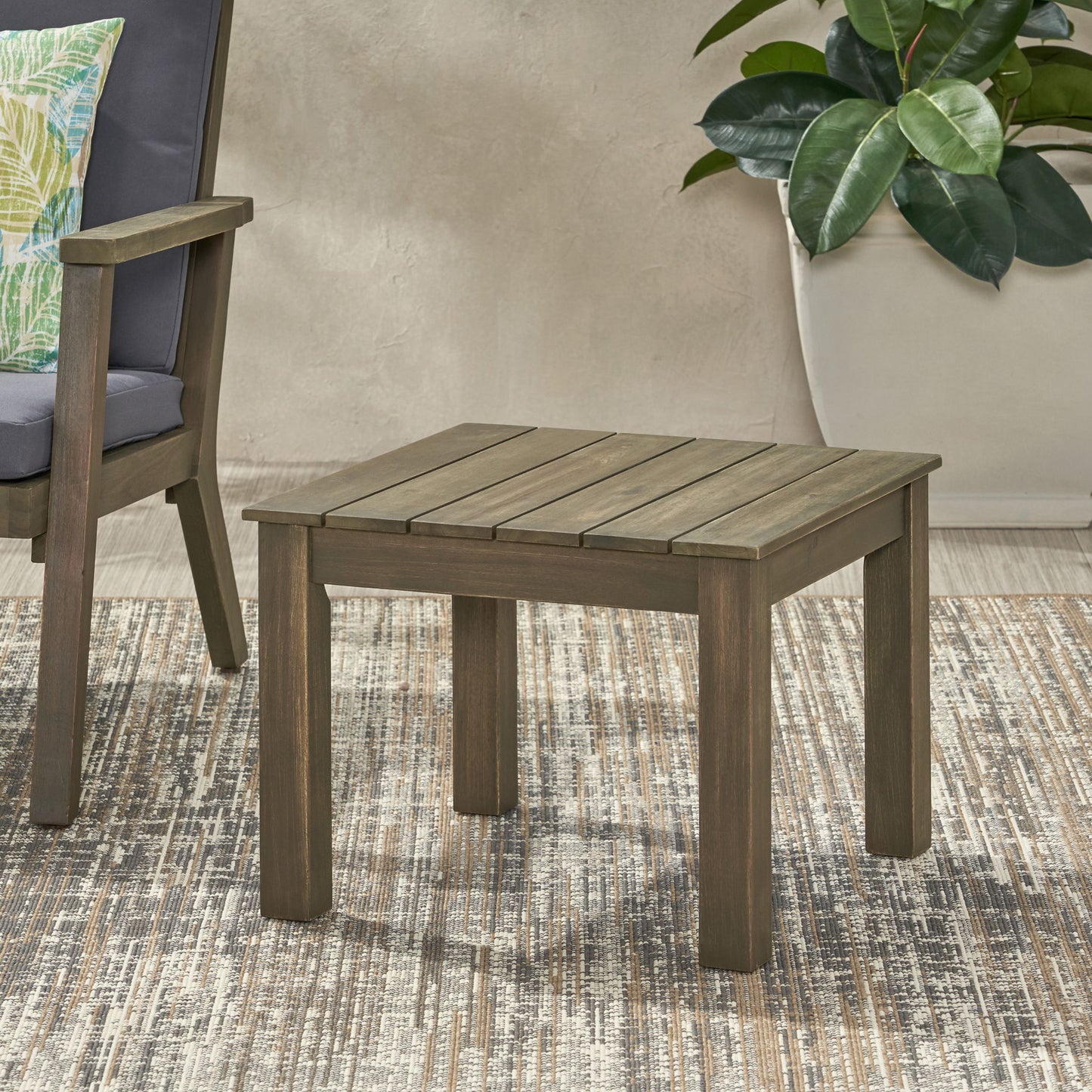 The Halcyon Outdoor Side Table with slat design sits on a patterned rug beside a cushioned chair, with a potted plant in the background.