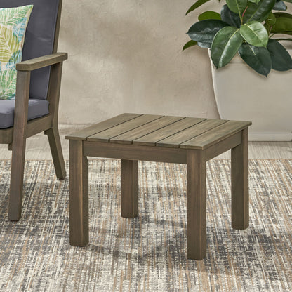 The Halcyon Outdoor Side Table with slat design sits on a patterned rug beside a cushioned chair, with a potted plant in the background.