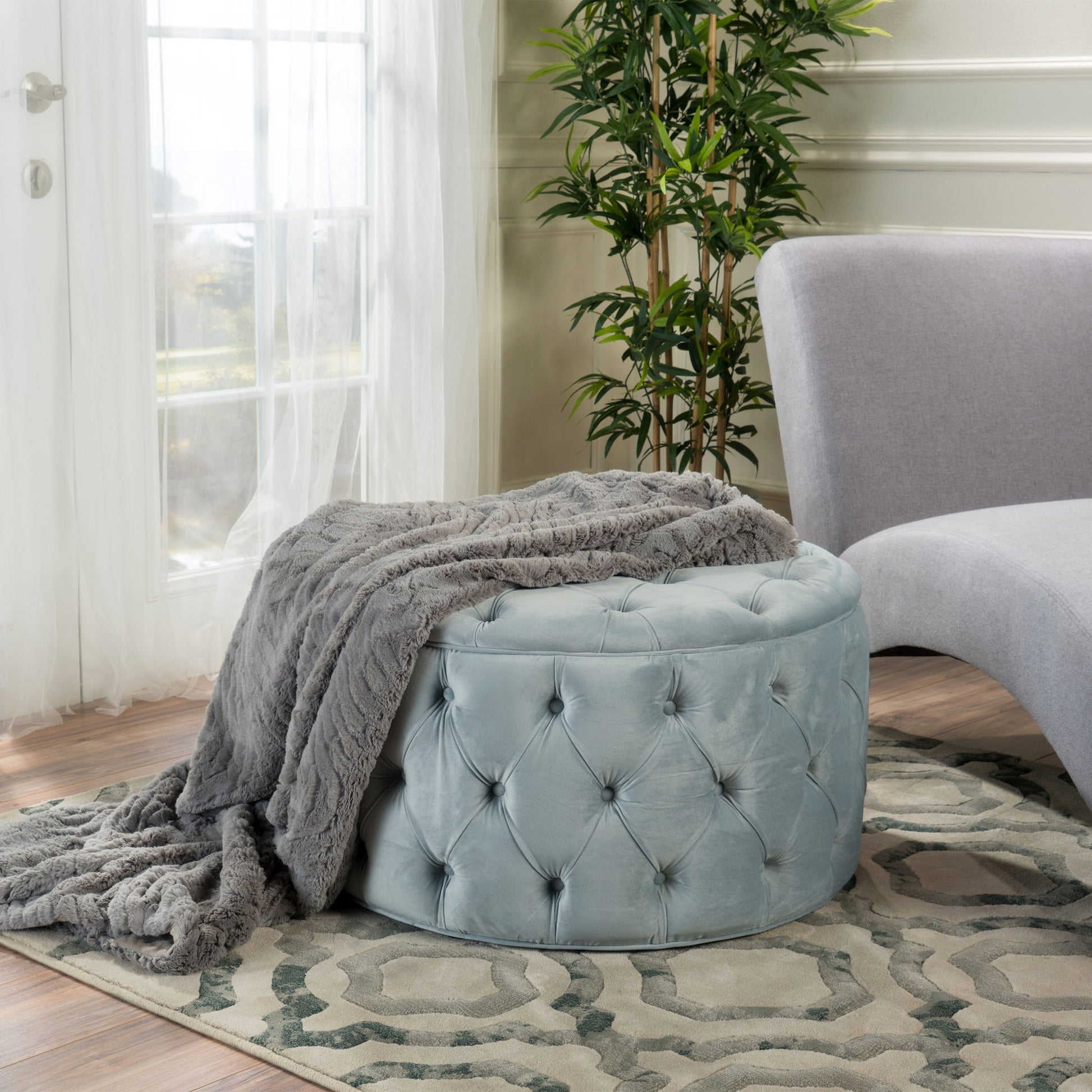 A Modern Glam Ottoman Velvet in light blue with a gray faux fur throw is placed on a patterned rug beside a light gray curved sofa, near a window with sheer curtains and a potted plant in the background.