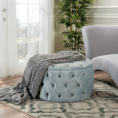 A Modern Glam Ottoman Velvet in light blue with a gray faux fur throw is placed on a patterned rug beside a light gray curved sofa, near a window with sheer curtains and a potted plant in the background.