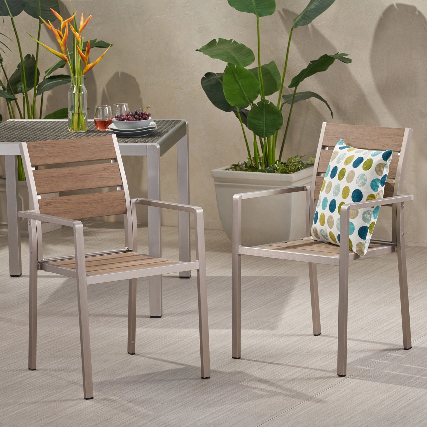 Two Dreamlan Outdoor Aluminum Dining Chairs with wood panels sit on a light floor; one has a colorful polka dot pillow. A nearby table holds drinks and grapes, while large green plants decorate the background.