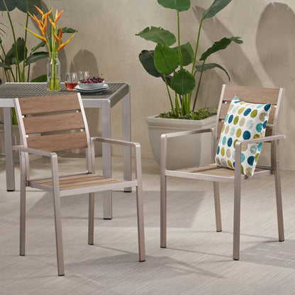 Two Dreamlan Outdoor Aluminum Dining Chairs with wood panels sit on a light floor; one has a colorful polka dot pillow. A nearby table holds drinks and grapes, while large green plants decorate the background.