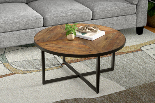 The Faro - Round Table, featuring a wooden top and black metal base, is placed on a patterned rug before a gray sofa. On the table sit a small potted plant, a book, and decorative accents.