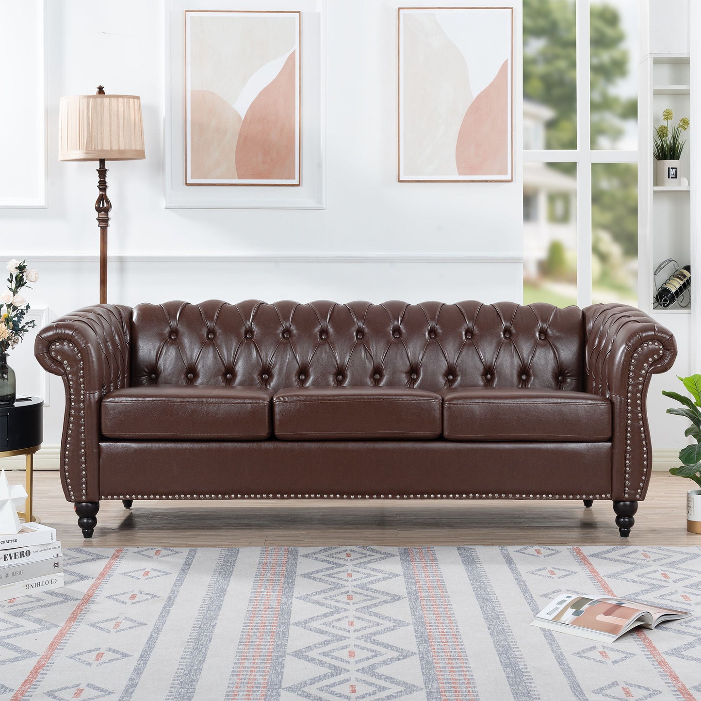 The Rolled Arm Chesterfield 3 Seater Sofa in dark brown leather with tufting and nailhead trim sits in a bright living room, accompanied by a lamp, potted plant, abstract wall art, and a patterned rug.