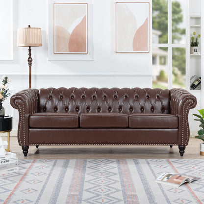 The Rolled Arm Chesterfield 3 Seater Sofa in dark brown leather with tufting and nailhead trim sits in a bright living room, accompanied by a lamp, potted plant, abstract wall art, and a patterned rug.