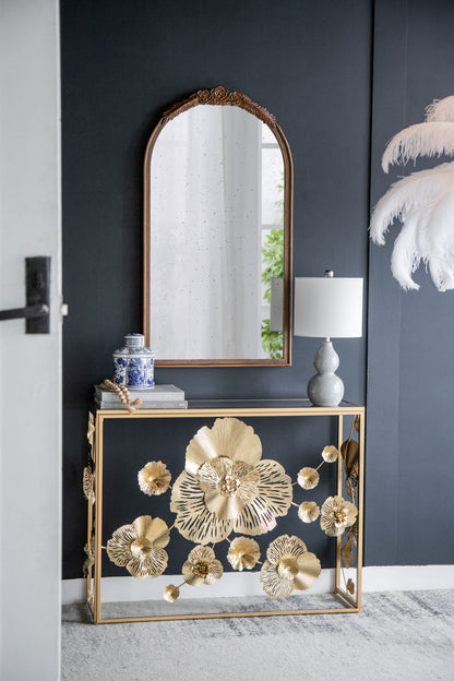 A gold console table topped with books, a blue-and-white vase, and a gray lamp stands against a dark wall. Above it hangs the Hand Carved Rose Antique Mirror Frame, Wood Arch Mirror Wall Decor For Living Room. White feathers sit to its right.