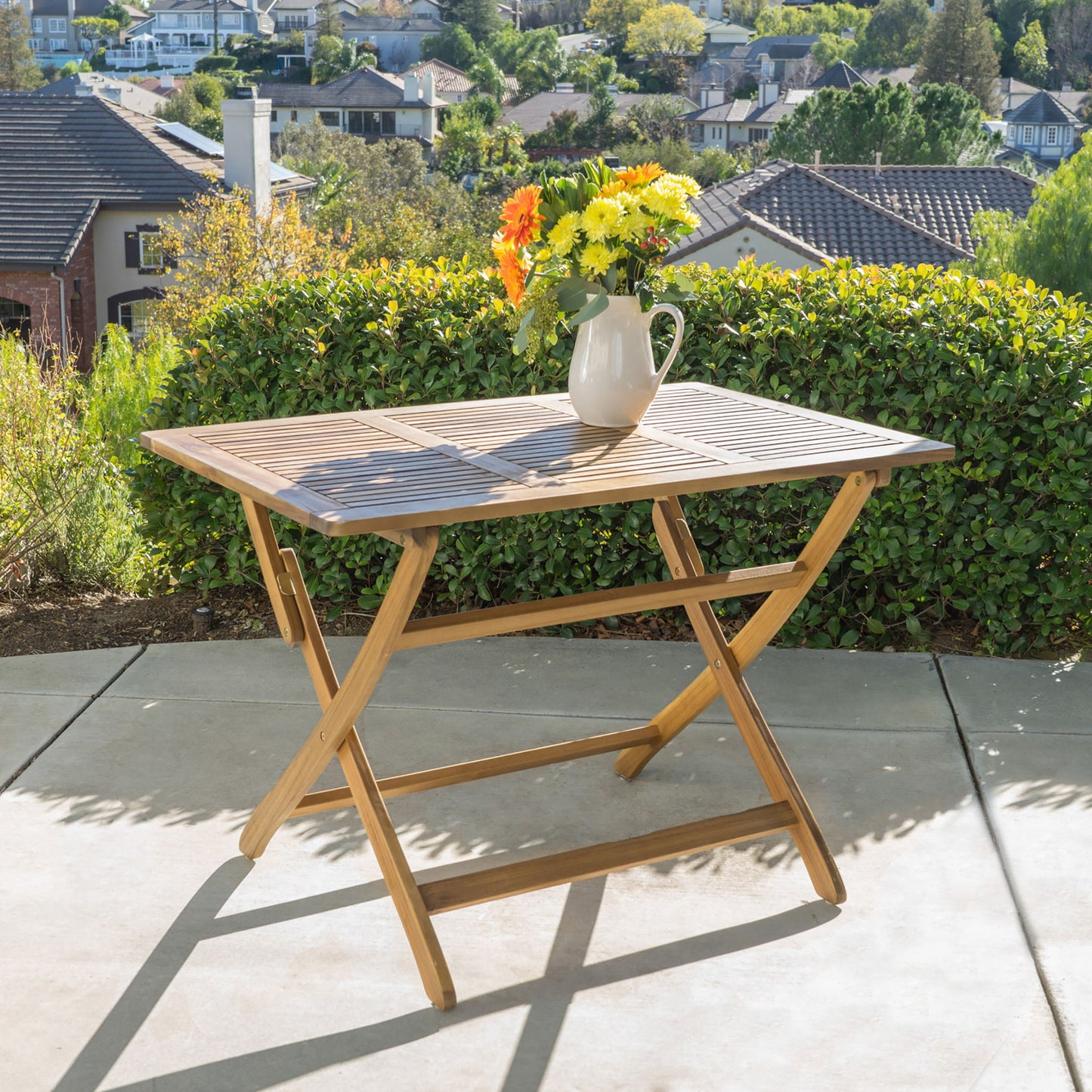 The Blissique Outdoor Foldable Wood Dining Table with a slat design and crossed legs stands on concrete, topped with a white pitcher of yellow and orange flowers. Green bushes, houses, and trees are visible in the background.