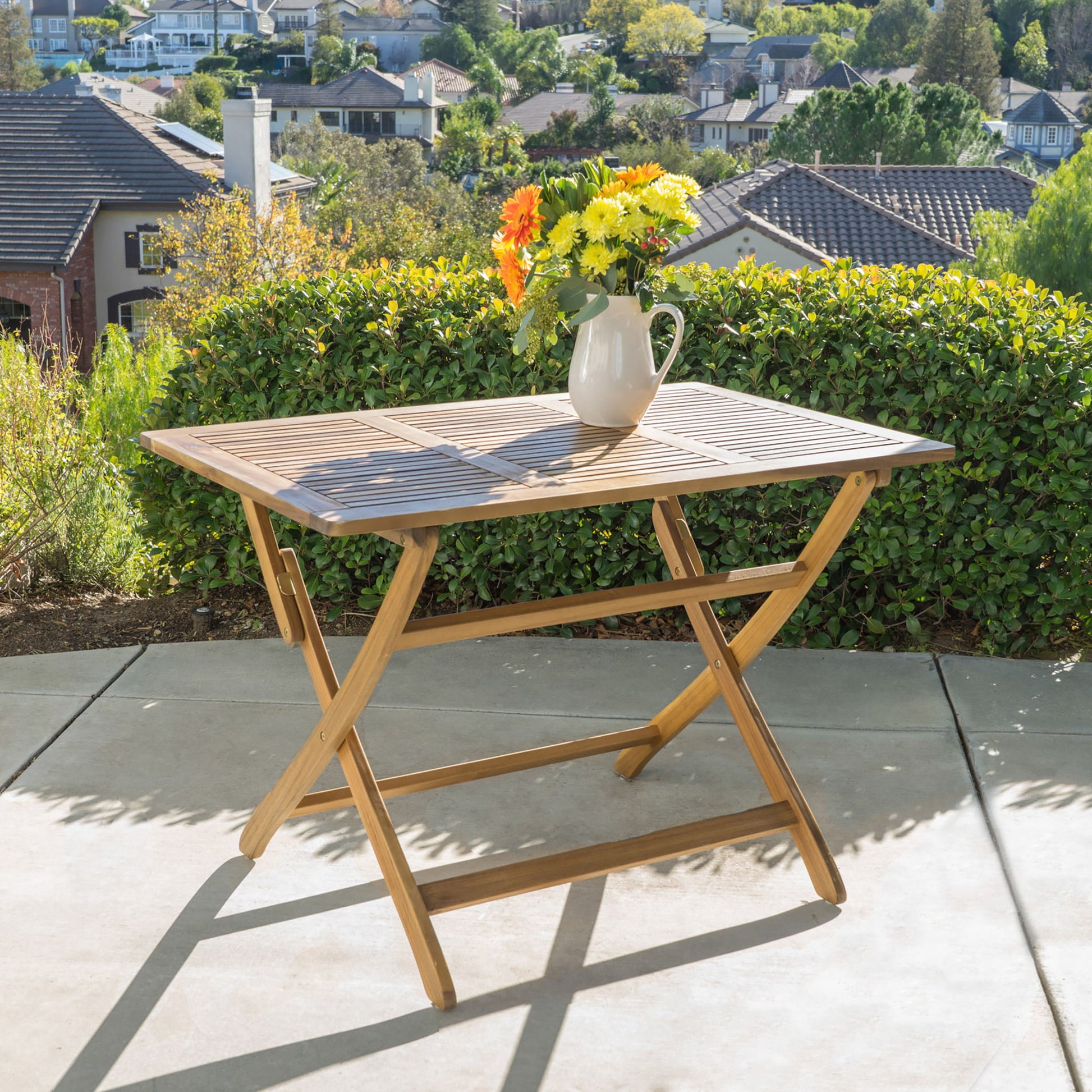 The Blissique Outdoor Foldable Wood Dining Table with a slat design and crossed legs stands on concrete, topped with a white pitcher of yellow and orange flowers. Green bushes, houses, and trees are visible in the background.