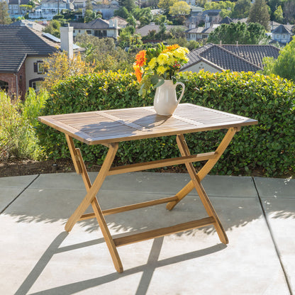 The Blissique Outdoor Foldable Wood Dining Table with a slat design and crossed legs stands on concrete, topped with a white pitcher of yellow and orange flowers. Green bushes, houses, and trees are visible in the background.