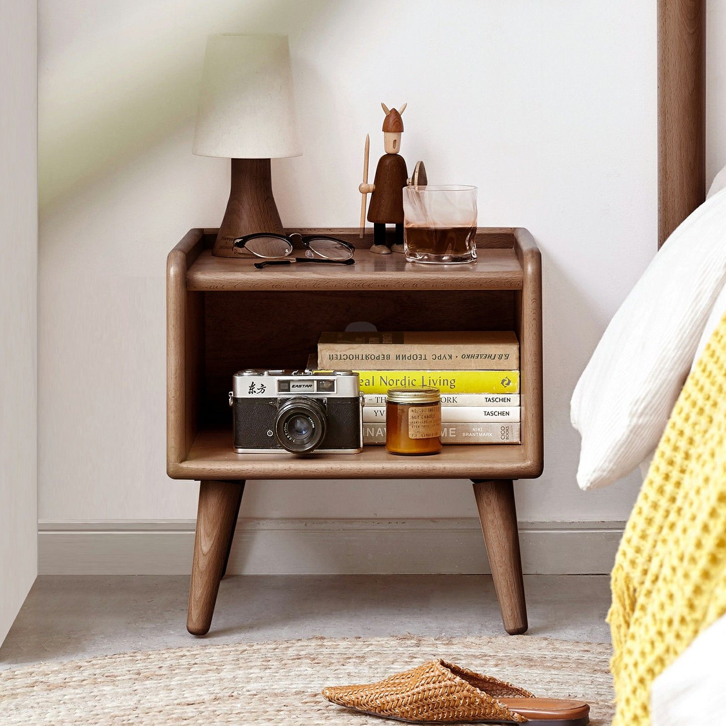 A Modern Simple Nightstand made of European Beech holds books, a vintage camera, candle, glasses, lamp, figurine, and iced coffee beside a bed with a white and yellow blanket.
