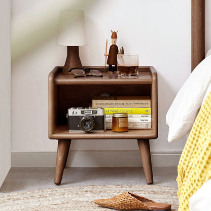 A Modern Simple Nightstand made of European Beech holds books, a vintage camera, candle, glasses, lamp, figurine, and iced coffee beside a bed with a white and yellow blanket.
