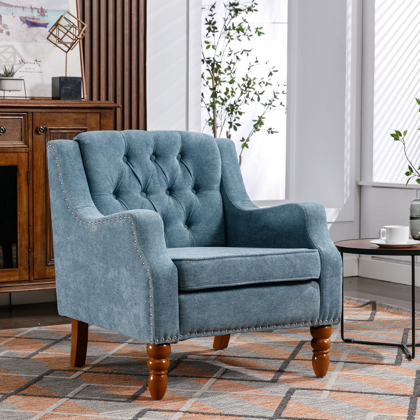 The Accent Chair, a button tufted upholstered armchair with vintage brass studs and footrest set, sits on a patterned rug beside a round table with tea, surrounded by sunlight, plants, and wooden furniture.