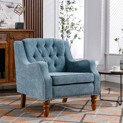 The Accent Chair, a button tufted upholstered armchair with vintage brass studs and footrest set, sits on a patterned rug beside a round table with tea, surrounded by sunlight, plants, and wooden furniture.