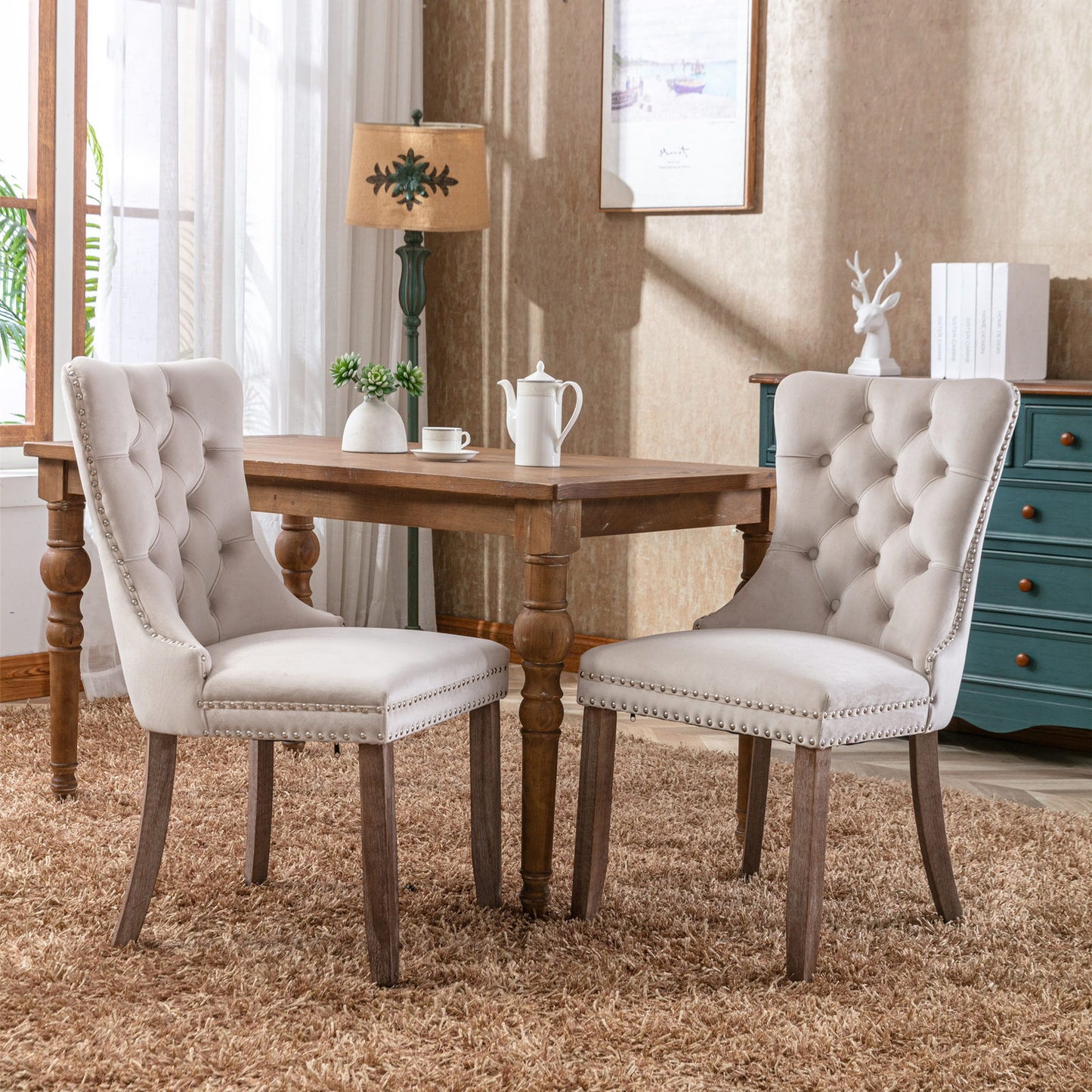 A set of two Nikki modern, high-end tufted velvet dining chairs with wood legs and nailhead trim sit at a square wooden table on a tan rug in a cozy, sunlit room with a lamp, dresser, and decorative accents in the background.