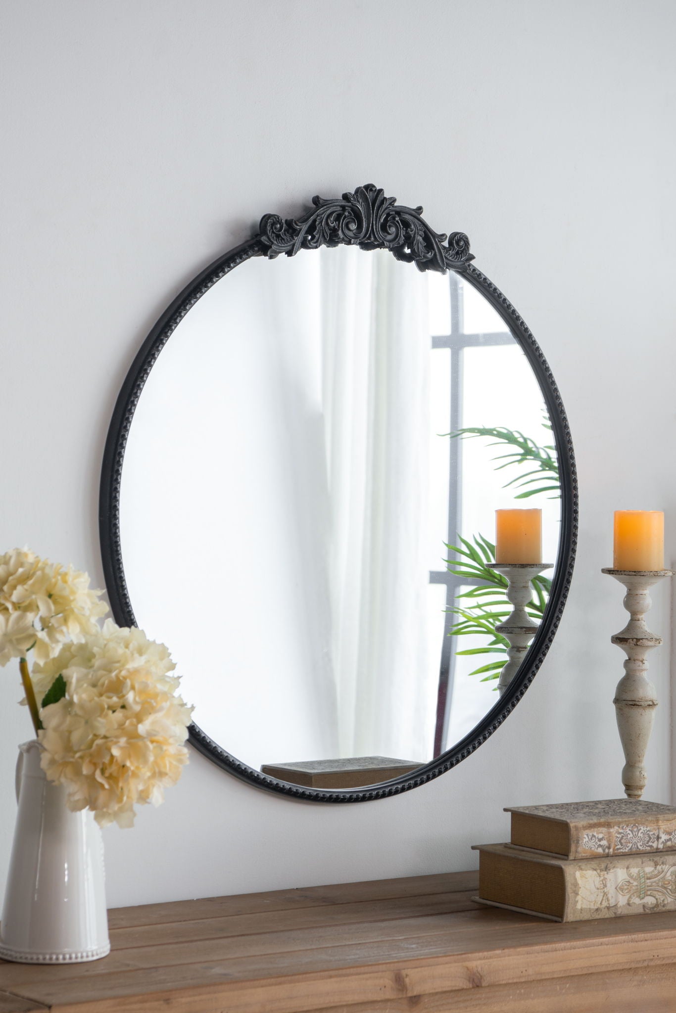 Classic Design Mirror with Baroque-inspired black frame reflects a window, white curtain, and plant. In front, a wood table holds stacked books, a white vase with cream flowers, and a lit candle on a tall holder. Ideal for bathrooms or entryways.