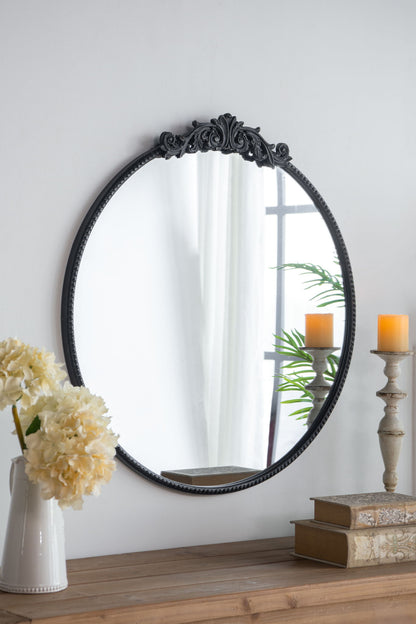 Classic Design Mirror with Baroque-inspired black frame reflects a window, white curtain, and plant. In front, a wood table holds stacked books, a white vase with cream flowers, and a lit candle on a tall holder. Ideal for bathrooms or entryways.