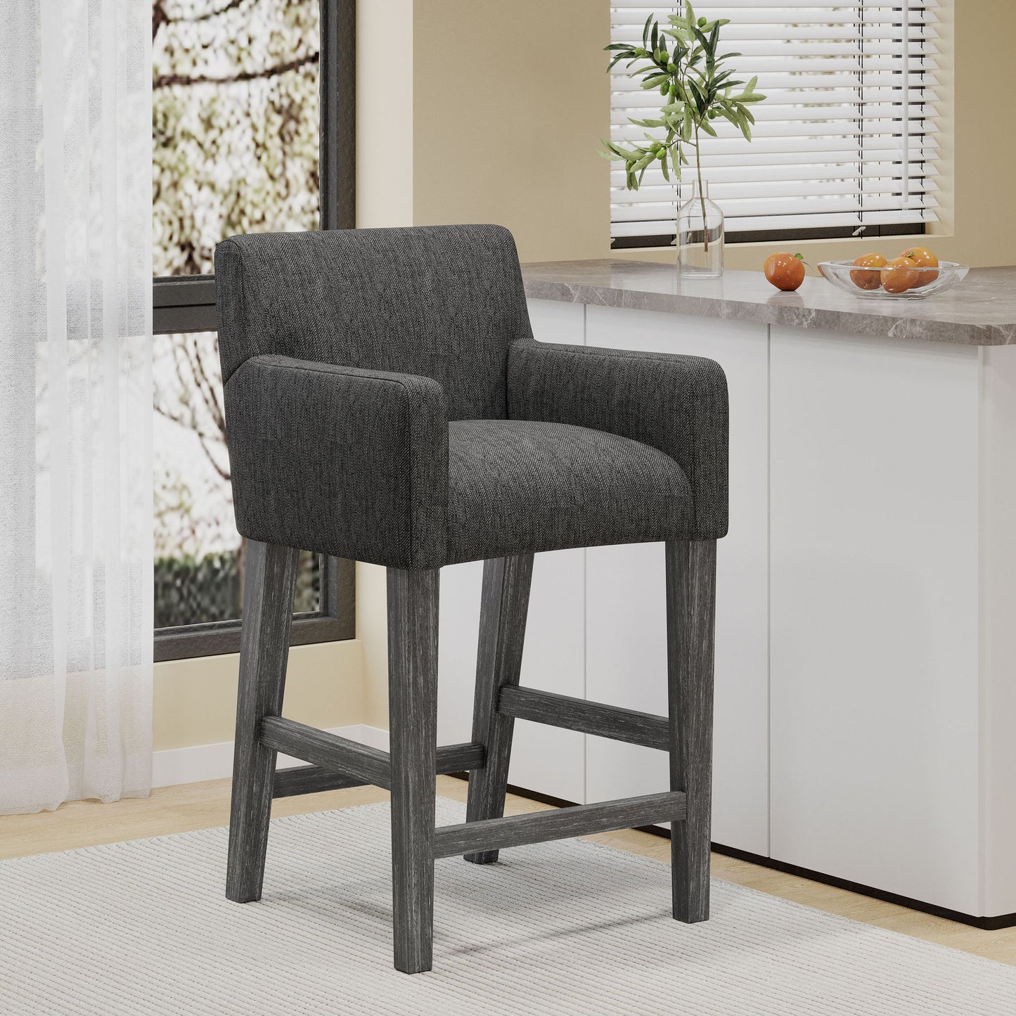 The Serenitia - Upholstered Counter Chair With Straight Arm features a dark gray upholstered seat and backrest, wooden legs, and a footrest, shown in a bright kitchen beside white cabinets and a window with blinds.