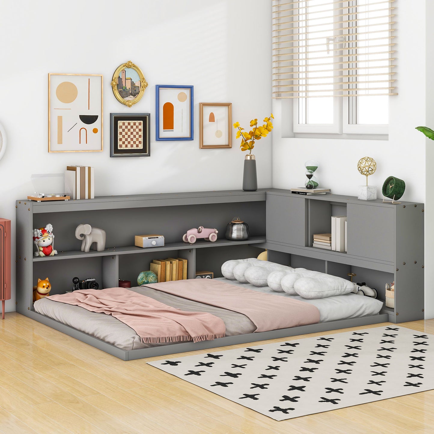 A cozy bedroom corner features the Floor Bed With L-Shaped Bookcases, Sliding Doors, Without Slats in grey, filled with books and decor. Pastel bedding, a black-and-white rug on wood floors, blinds, and framed art complete the look.