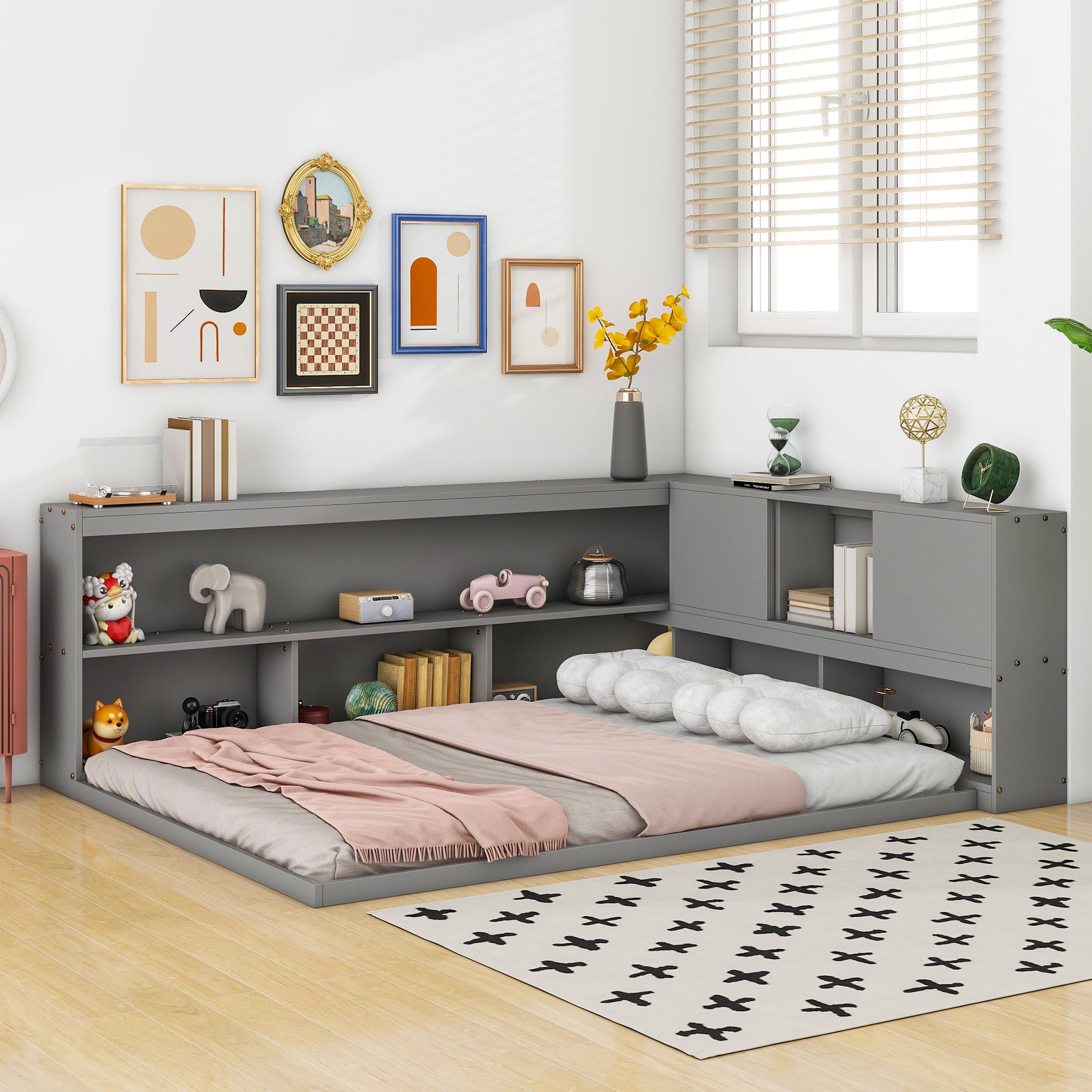 A cozy bedroom corner features the Floor Bed With L-Shaped Bookcases, Sliding Doors, Without Slats in grey, filled with books and decor. Pastel bedding, a black-and-white rug on wood floors, blinds, and framed art complete the look.