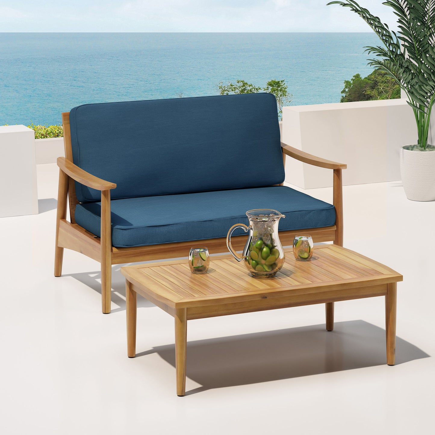 The Outdoor Acacia Wood Loveseat Set With Coffee Table features a wooden loveseat with blue cushions and a matching table on a patio overlooking the ocean, with citrus water in a glass pitcher, two glasses, and a potted plant nearby.