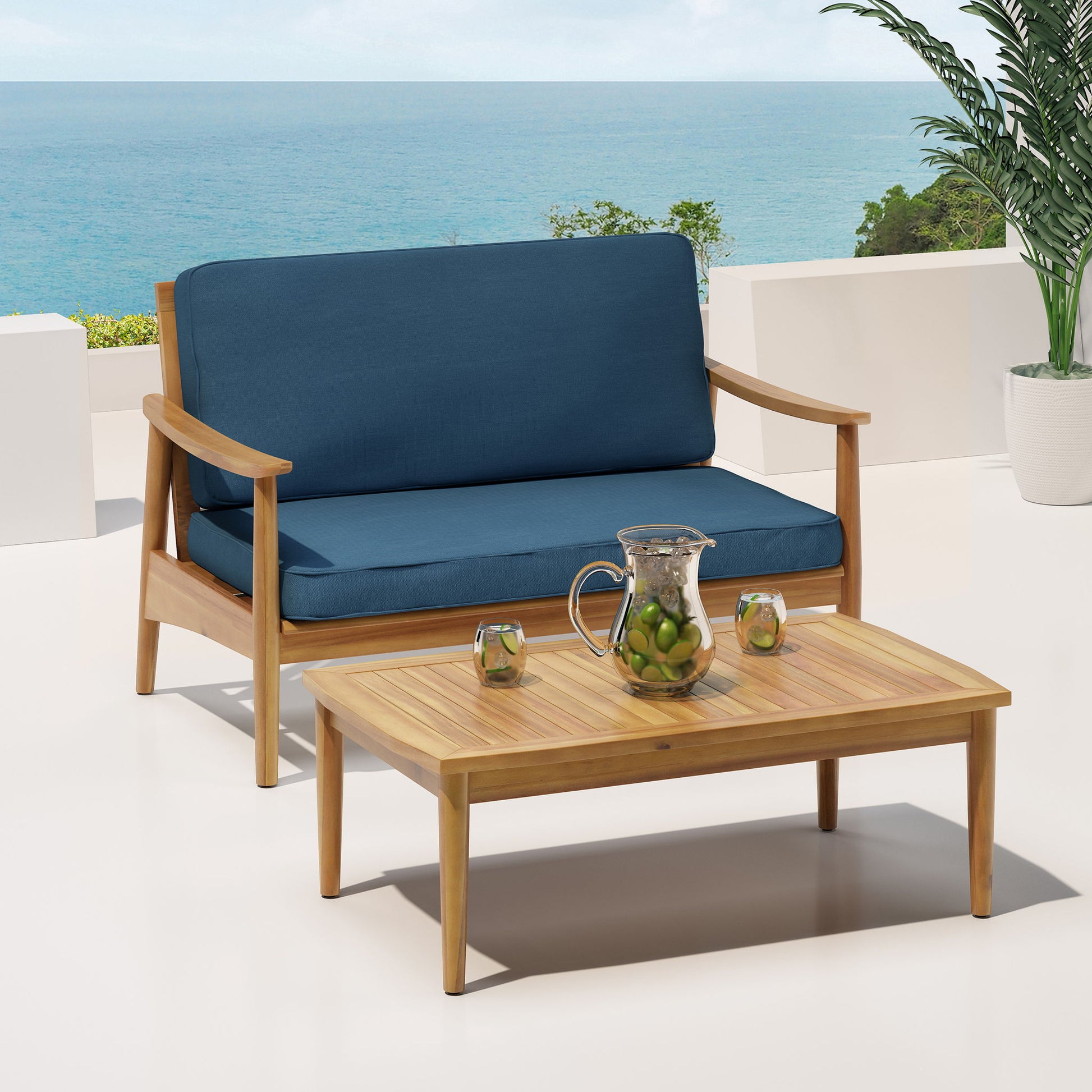 The Outdoor Acacia Wood Loveseat Set With Coffee Table features a wooden loveseat with blue cushions and a matching table on a patio overlooking the ocean, with citrus water in a glass pitcher, two glasses, and a potted plant nearby.