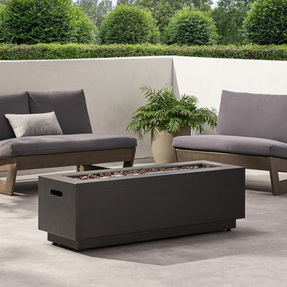 Modern outdoor patio with gray cushioned seating, a Modern Rectangular Iron Fire Pit 50,000 Btu with lava rocks, gray pillow, potted plant, and trimmed green bushes in the background.