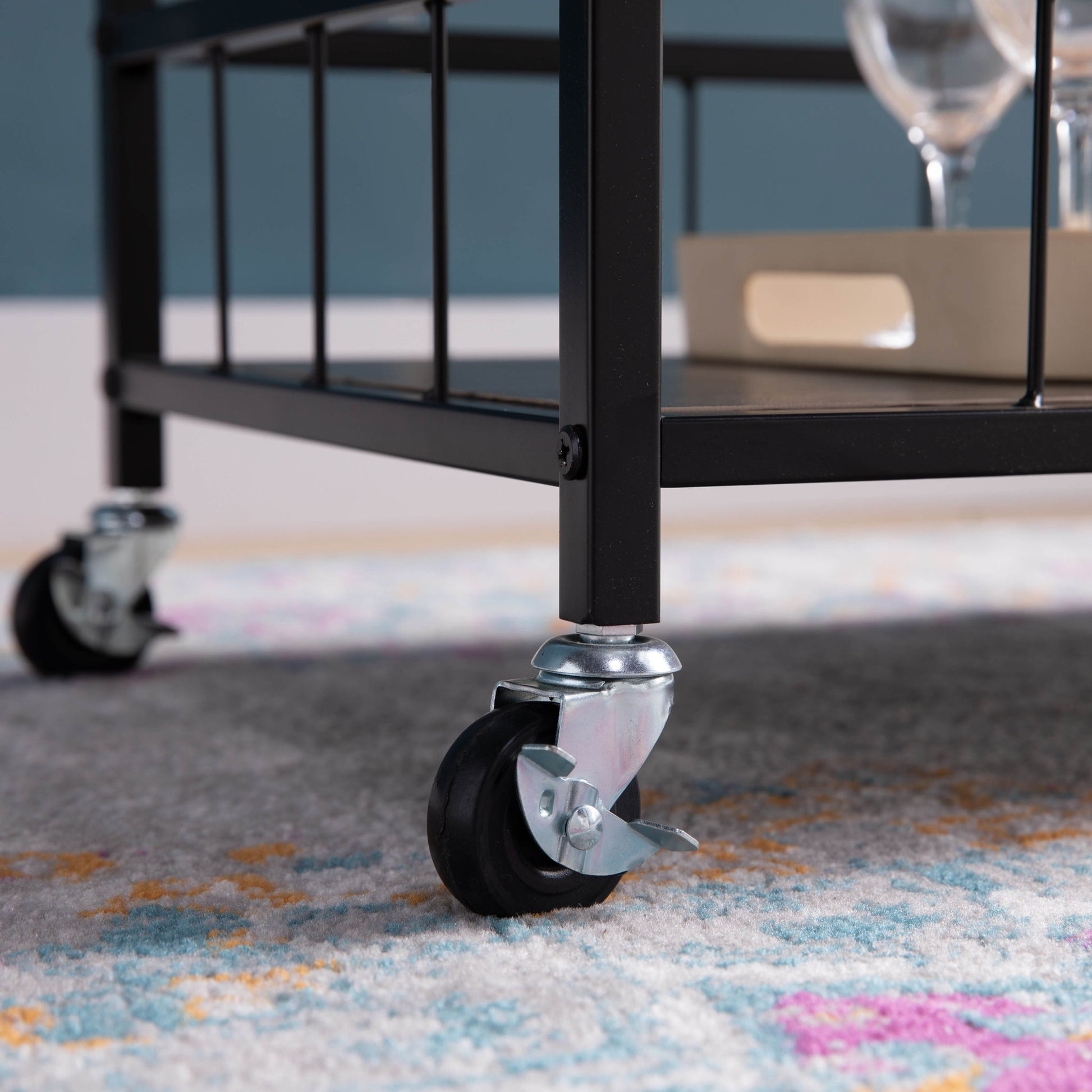 Close-up of the black metal leg and swivel caster wheel of the Rolling Kitchen Cart With Storage And Four Wine Bottle Rack on patterned carpet, with a lower shelf tray holding glassware visible in the background.