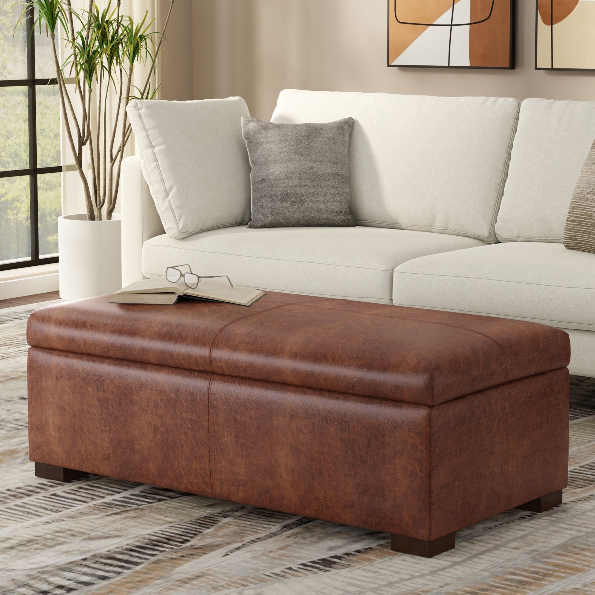 The Gabbie Coffee Table Upholstered Storage Ottoman with eyeglasses on top sits in front of a light-colored sofa with gray and white pillows in a modern living room. Abstract art hangs above the sofa.
