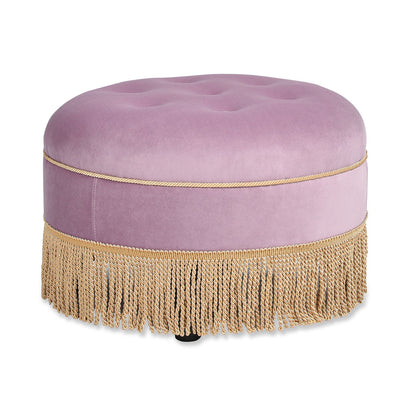 The Yolanda Round Upholstered Accent Ottoman features soft lavender fabric, gold trim, and decorative gold fringe at the base for a plush, elegant look.