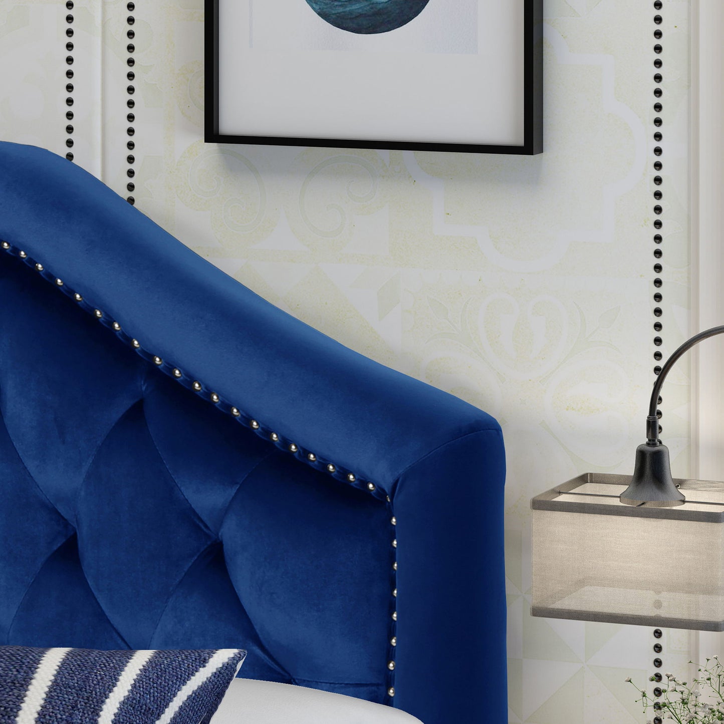 A close-up of the Kaarina - Contemporary Tufted Fabric Headboard in blue velvet with silver stud trim beside a wall-mounted lamp and framed abstract art on a patterned wall. A striped pillow appears at the bottom.