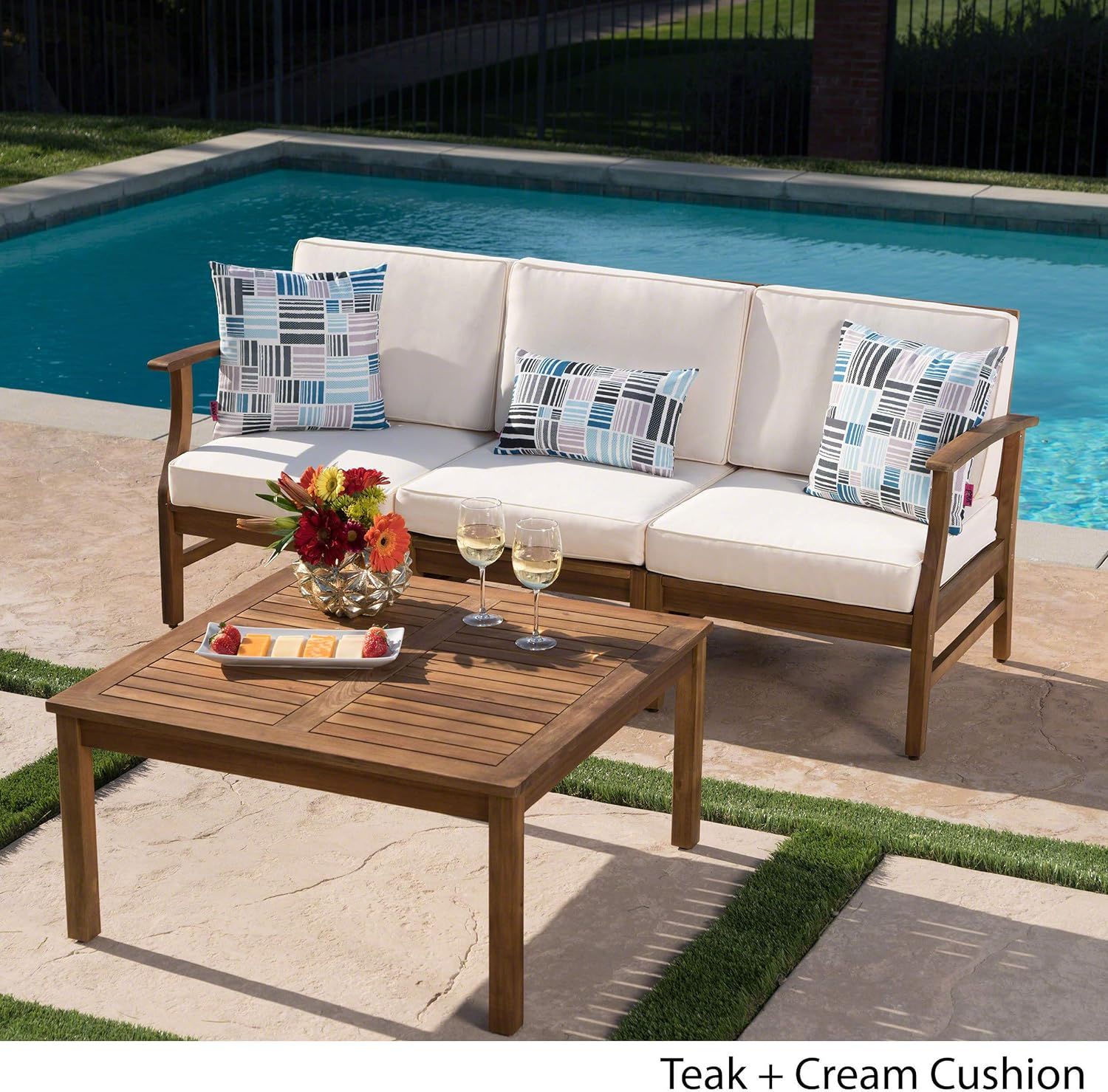 The Perla - 3 Seater Sofa and Table Set, with cream cushions and patterned pillows, is arranged by a poolside. The matching table displays flowers, three glasses of white wine, and a small plate of food.