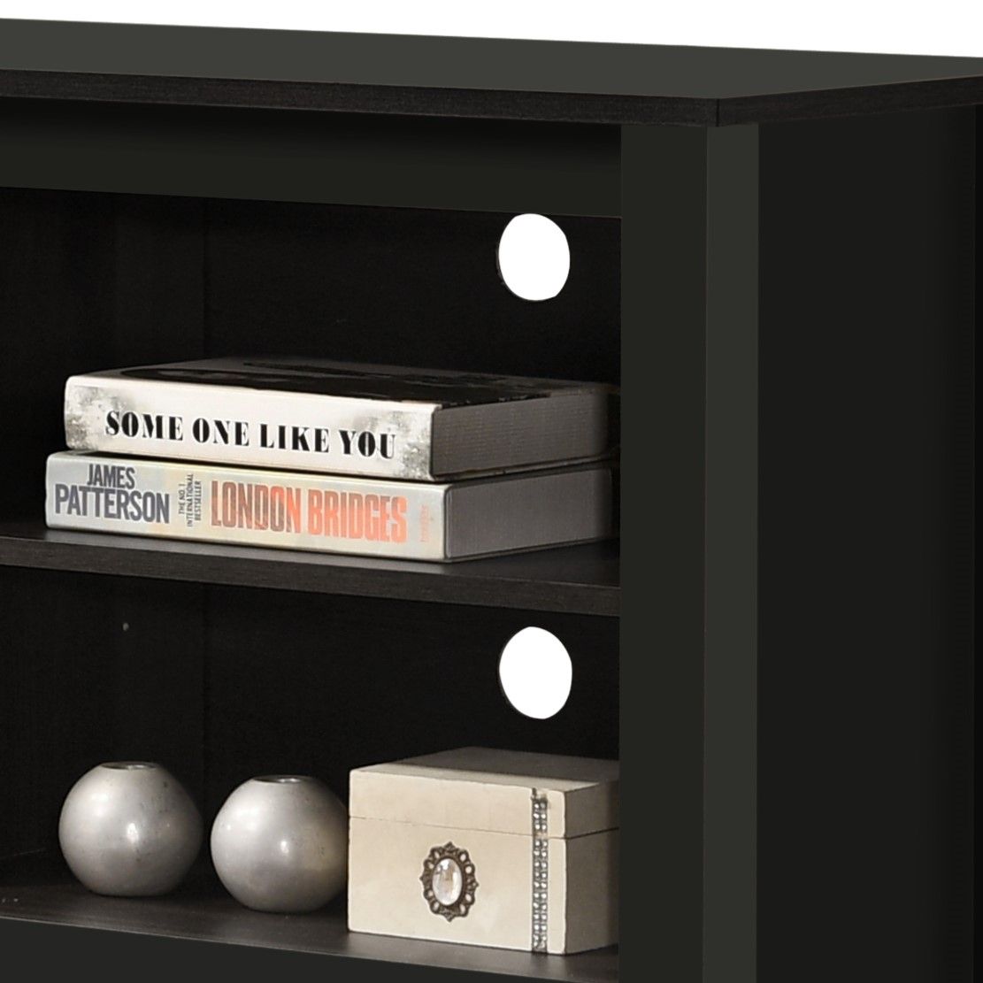 A close-up of the Benito TV Stand with open shelves and cable management. The black stand features two shelves: the top holds three books, while the bottom displays round décor items and a beige box. Two circular cord holes are visible at the back.