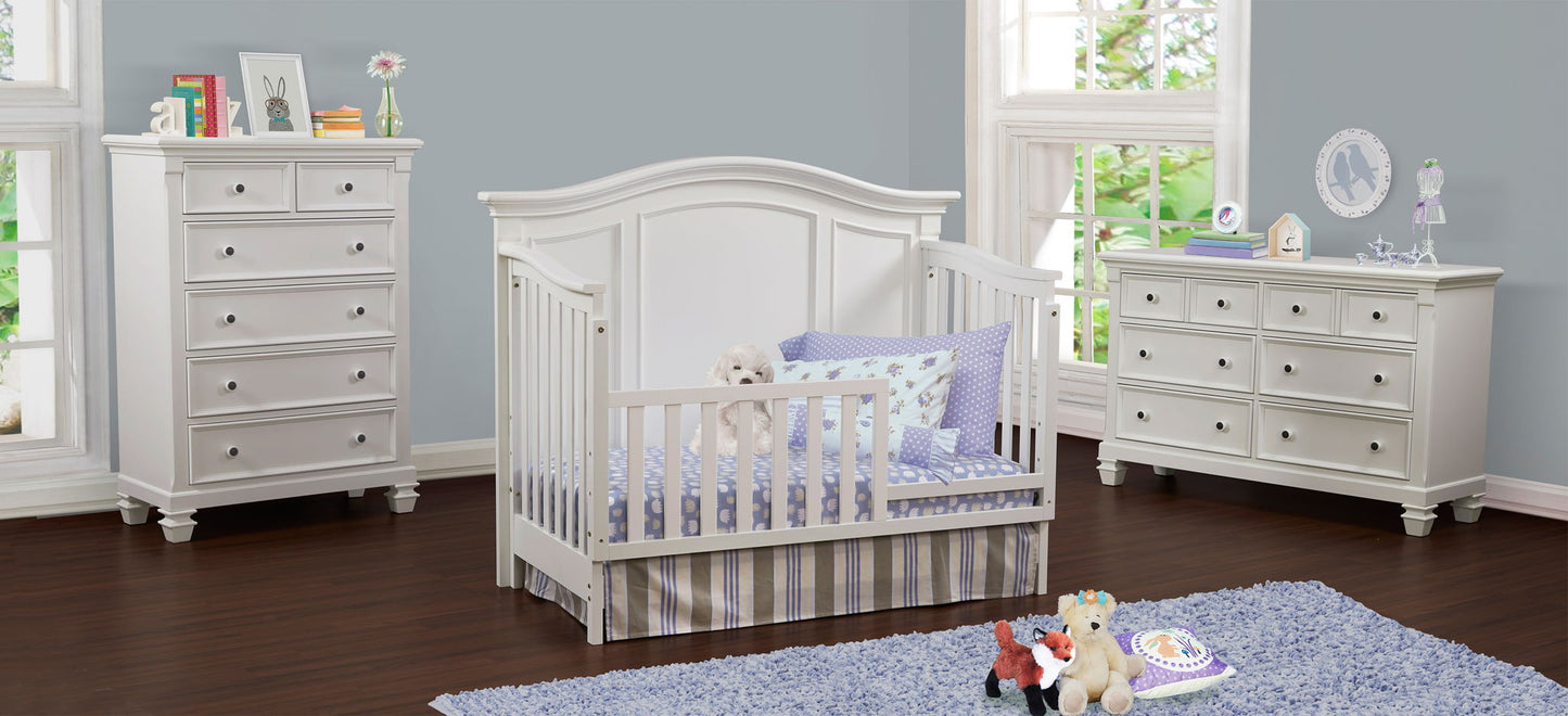 A nursery with a white crib, stuffed animals, purple bedding, and a Glendale - 6 Drawer Chest. The room features light blue walls, large windows, hardwood floors, and a blue rug scattered with toys.