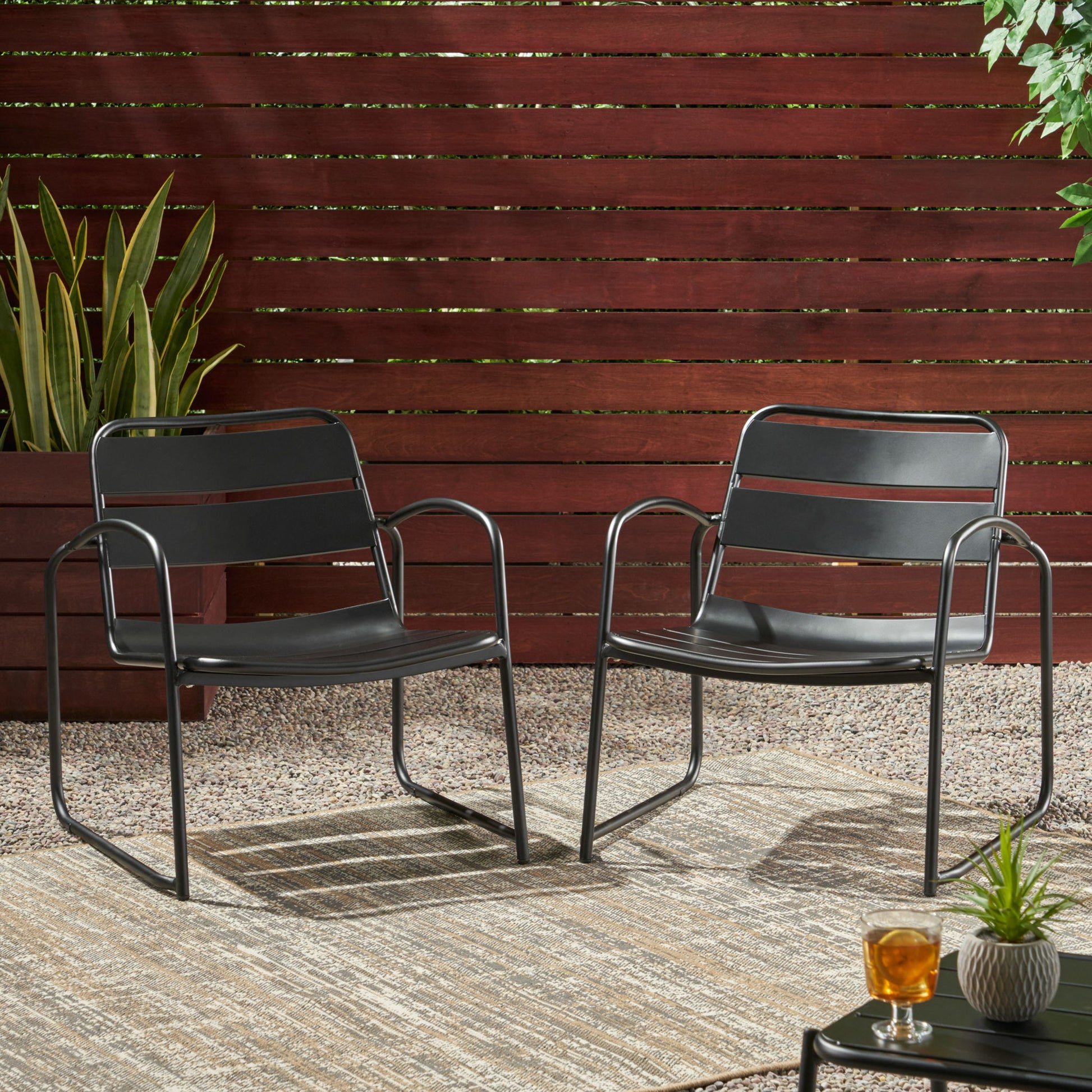 Two Conrad outdoor chairs in modern black metal sit on a patterned rug in a gravel patio, framed by a wooden fence and greenery. A small table with a glass of tea is placed in the foreground.