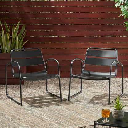 Two Conrad outdoor chairs in modern black metal sit on a patterned rug in a gravel patio, framed by a wooden fence and greenery. A small table with a glass of tea is placed in the foreground.