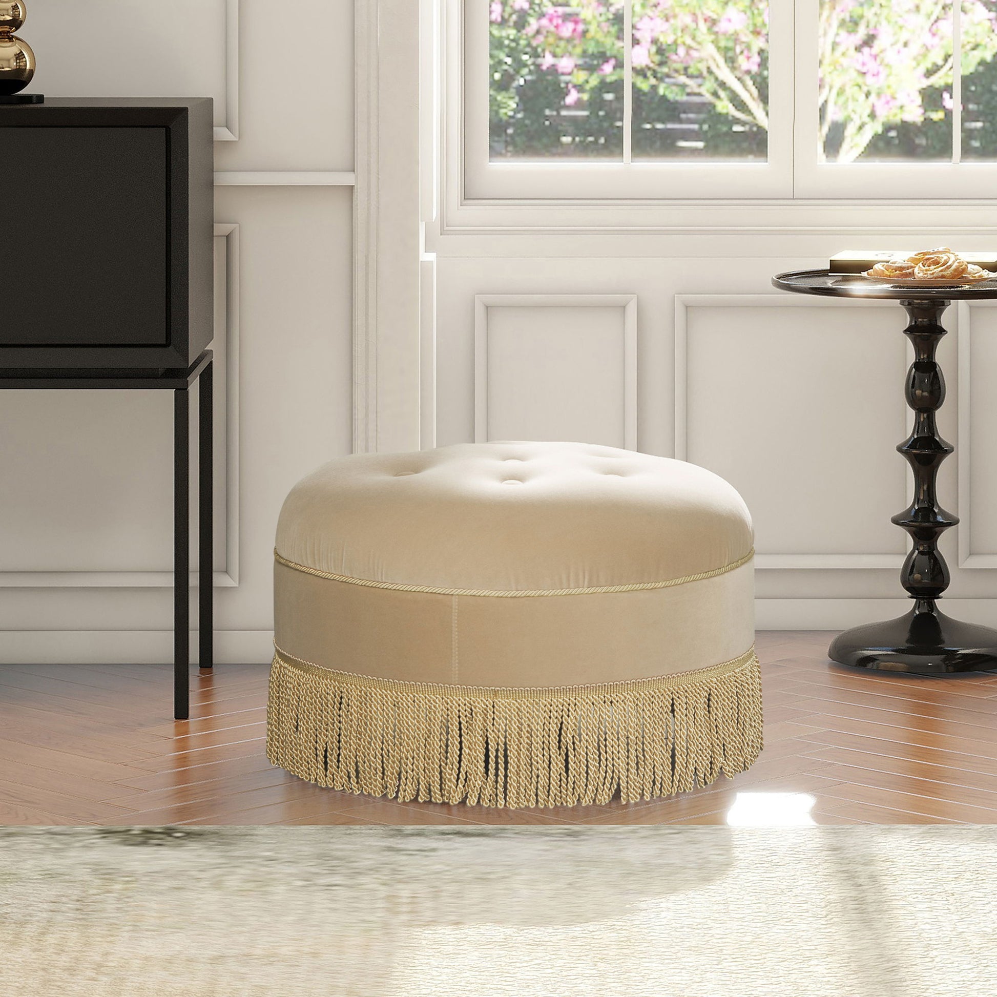 The Yolanda - Round Upholstered Accent Ottoman, in beige with gold fringe trim, sits on a wooden floor in a bright room with white paneled walls, a black side table, and large windows overlooking greenery and flowers.