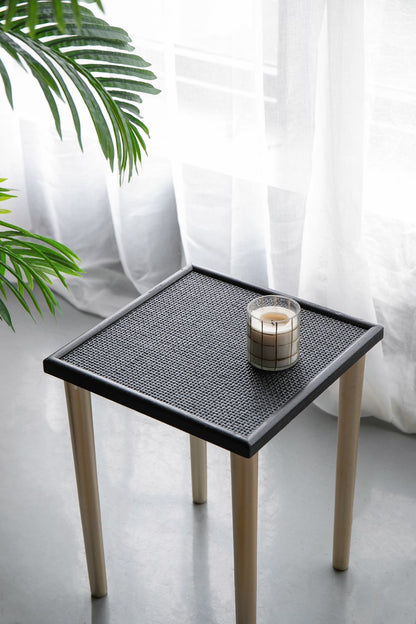 A Side Tables (Set of 3) - Black / Gold holds a lit candle in a glass holder. Sheer white curtains and green plant leaves can be seen in the background.