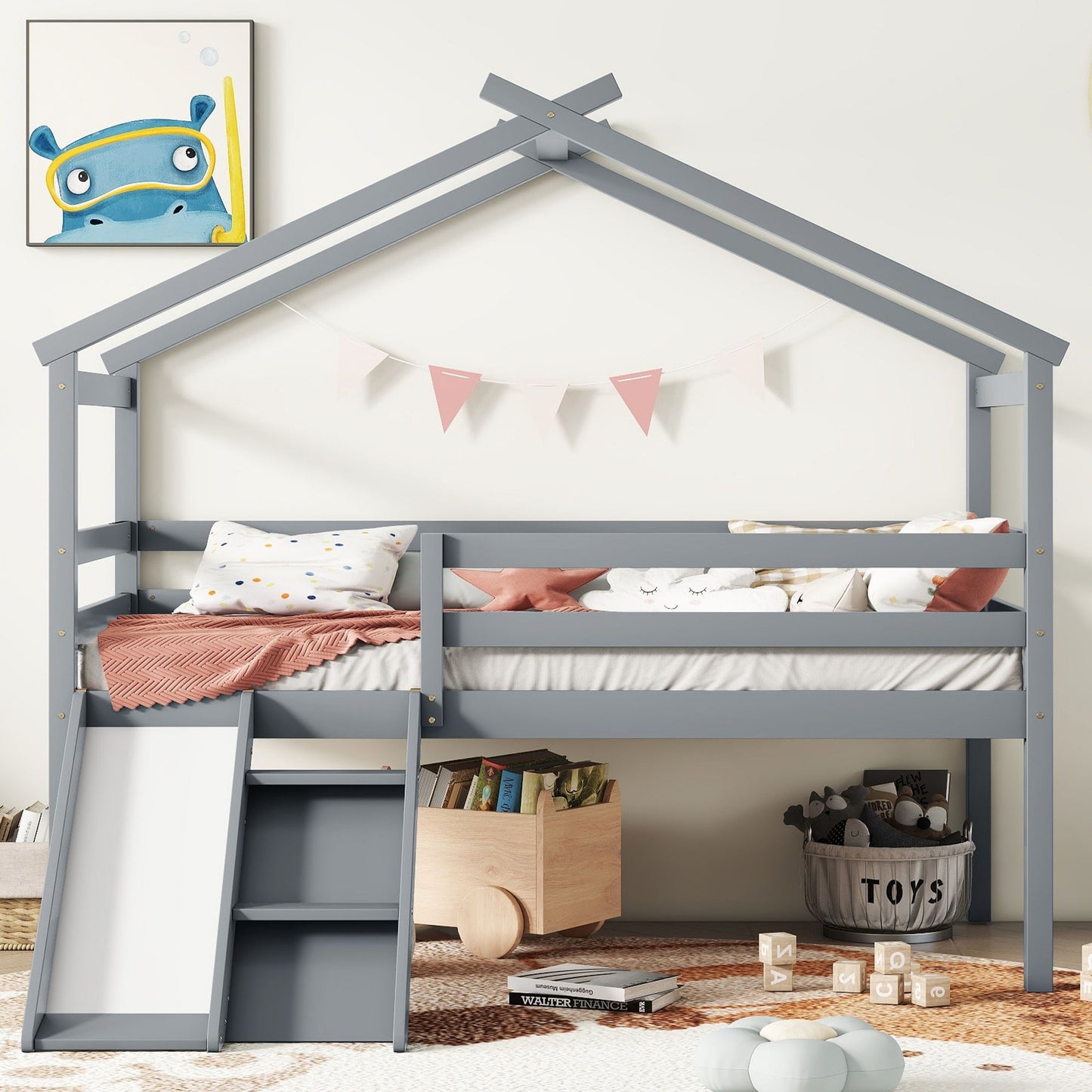 A gray Low Loft House Bed With Slide, Ladder, Safety Guardrails, and a house roof frame is decorated with pink bunting, pillows, and bedding. A wooden toy box with toys sits on a striped rug below, with framed cartoon art on the wall.