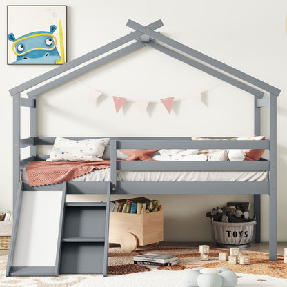 A gray Low Loft House Bed With Slide, Ladder, Safety Guardrails, and a house roof frame is decorated with pink bunting, pillows, and bedding. A wooden toy box with toys sits on a striped rug below, with framed cartoon art on the wall.