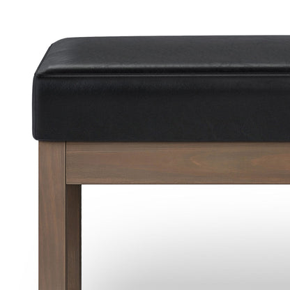A close-up of the Milltown - Upholstered Ottoman Bench, featuring a black cushioned seat and wooden legs, set against a plain white background.