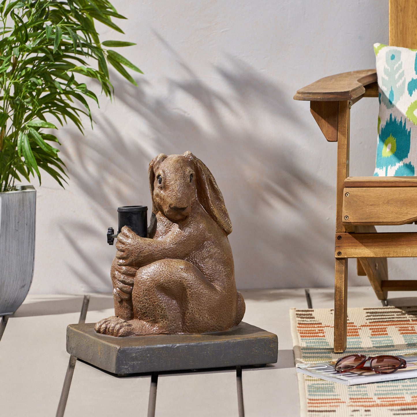 A light brown Hand Crafted Rabbit Umbrella Base with a playful design is placed on a patio deck beside a potted plant, a wooden chair featuring a colorful cushion, and a patterned rug.